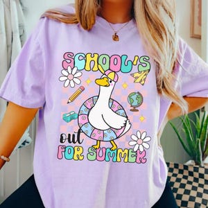 Pu&ograve; includere: T-shirt lilla chiaro con grafica di un'oca dei cartoni animati con cappello e salvagente. Il testo "SCHOOL'S OUT FOR SUMMER" &egrave; in lettere colorate, con illustrazioni a tema scolastico.