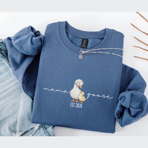 May include: A blue sweatshirt with the words "mama goose" in cursive and a graphic of a mother goose and gosling. The sweatshirt also has the text "EST. 2026". A silver necklace is draped across the top of the sweatshirt.