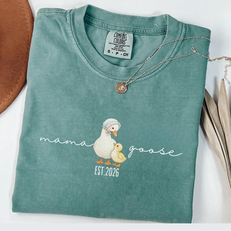 May include: A teal Comfort Colors t-shirt with a Mama Goose graphic and the text "mama goose EST. 2026". The graphic features a mother goose and a baby goose. A silver necklace with a pendant is on the shirt.
