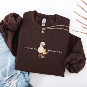 May include: A dark brown sweatshirt with the words "mama goose" and a graphic of a mother goose and gosling. The sweatshirt also has the text "EST. 2026". A silver necklace with a pendant is draped across the sweatshirt.