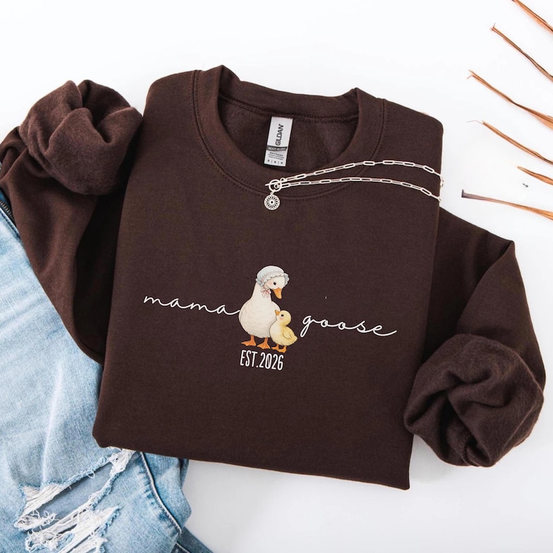 May include: A dark brown sweatshirt with the words "mama goose" and a graphic of a mother goose and gosling. The sweatshirt also has the text "EST. 2026". A silver necklace with a pendant is draped across the sweatshirt.