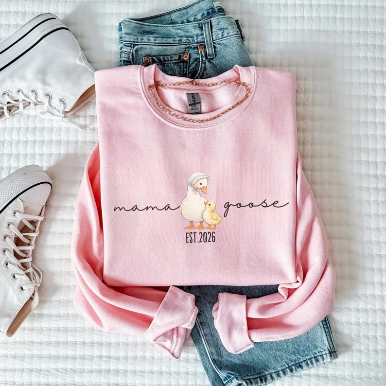 May include: A light pink sweatshirt with the words "mama goose" in a script font, along with an illustration of a goose and a gosling. The text "EST. 2026" is below the illustration. The sweatshirt is paired with blue jeans and white sneakers.