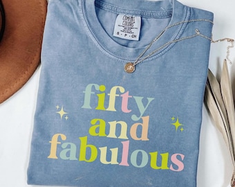 May include: A blue Comfort Colors t-shirt with the words "fifty and fabulous" in colorful letters. The shirt has a crew neck and short sleeves. A gold necklace with a pendant is draped across the shirt.