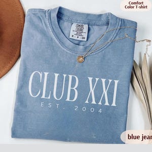 May include: A blue jean Comfort Colors t-shirt with the text "CLUB XXI EST. 2004" in white. The shirt has a crew neck and a small tag on the collar. A gold necklace with a pendant is draped across the shirt.