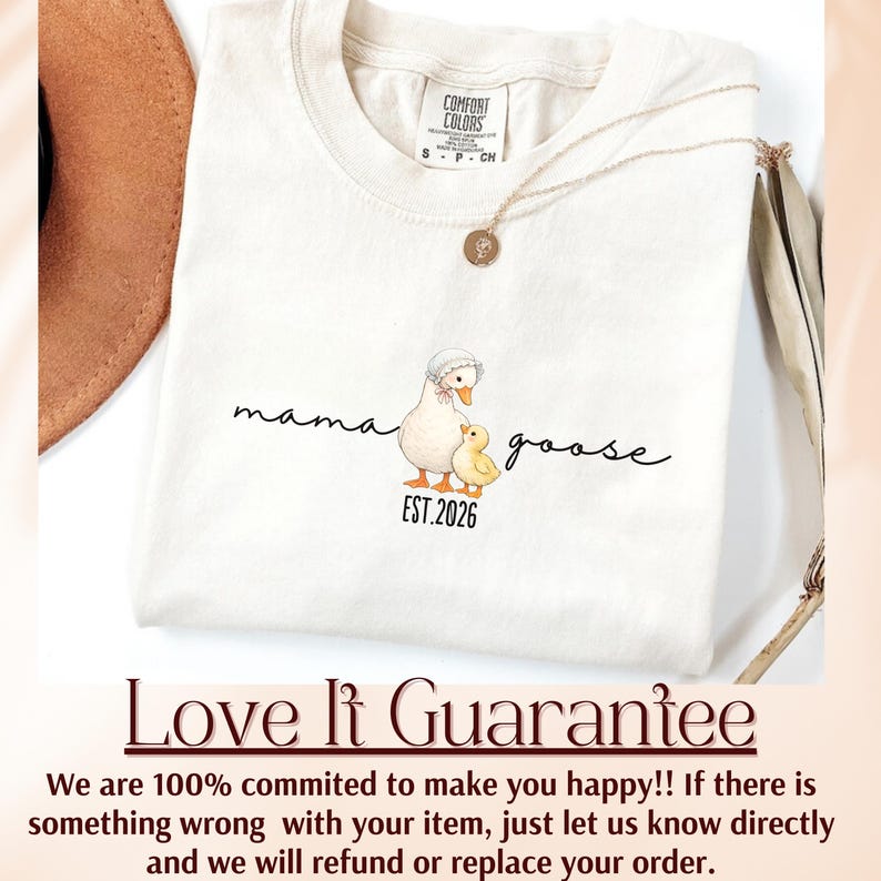 May include: Cream-colored t-shirt with a Mama Goose graphic. The design features a goose wearing a bonnet and a yellow chick, with the text "mama goose" and "EST.2026" in a script font. A gold necklace is draped across the shirt.
