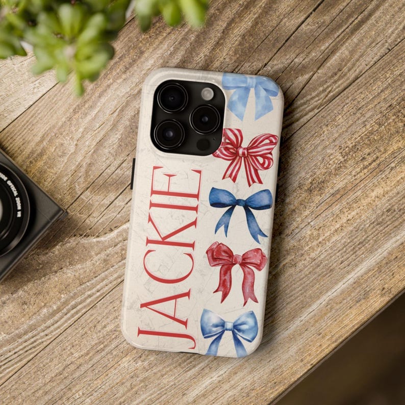 Puede incluir: Una funda de tel&eacute;fono con fondo crema y el nombre "JACKIE" en letras rojas. La funda presenta lazos de acuarela en rojo y azul. La funda del tel&eacute;fono est&aacute; sobre una superficie de madera.
