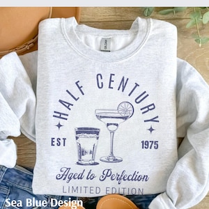 May include: A light gray sweatshirt with a navy blue graphic that reads "HALF CENTURY" above a cocktail and a shot glass. The text "Aged to Perfection" and "LIMITED EDITION" are below the image. The year "1975" is also printed on the sweatshirt.