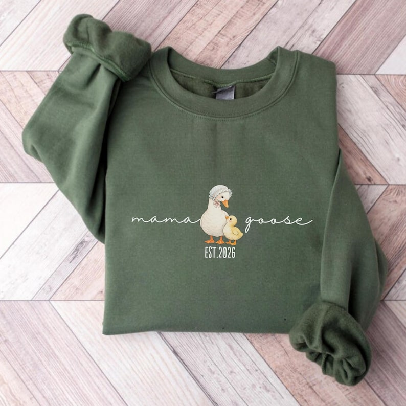 May include: An olive green sweatshirt featuring a graphic of a mother goose and a baby chick. The words "mama goose" are written in cursive above the illustration, with "EST. 2026" below. The sweatshirt has long sleeves.