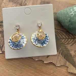 May include: Dangle earrings with miniature teacups and saucers. The teacups and saucers are white with blue floral patterns and gold trim. Each earring has a clear, round charm and a sparkling, clear crystal stud.