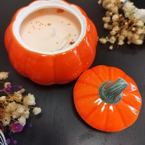 Könnte beinhalten: Ein leuchtend orangefarbener Keramik-Kerzenbehälter in Kürbisform mit passendem Deckel und grünem Stiel. Das Kerzenwachs ist hellbeige und mit kleinen goldenen sternförmigen Glitzerelementen, winzigen bunten Blättern und einem Kristall verziert.