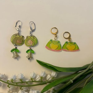 Könnte beinhalten: Zwei Paar handgefertigte Ohrringe. Ein Paar zeigt grüne Froschdesigns mit gelben Herzen und rosa Akzenten. Das andere Paar hat grüne Froschdesigns mit Spiraldetails. Beide Paare haben silber- und goldfarbene Hardware.