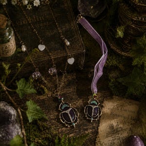Könnte beinhalten: Eine goldfarbene Halskette mit einem dunkelvioletten Käferanhänger mit klaren Kristallen, kleinen weißen Herzanhängern und Perlen, sowie ein passendes violettes Samtband-Halsband mit einem ähnlichen Käferanhänger mit türkisfarbenen Akzenten.