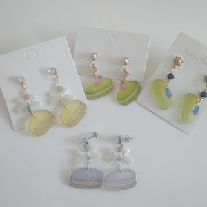 Könnte beinhalten: Eine Auswahl an Ohrhängern mit bunten Macaron-Designs. Die Ohrringe sind in verschiedenen Farben erhältlich, darunter Gelb, Grün und Lila, mit dekorativen Schleifen und Kristallakzenten. Die Ohrringe werden auf weißen Ohrringkarten präsentiert.