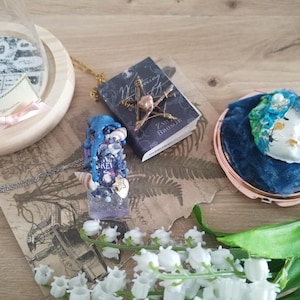 Könnte beinhalten: Eine Auswahl an handgefertigtem Schmuck und Accessoires, darunter ein Miniatur-Buchanhänger mit einer Goldkette, ein Resin-Anhänger mit dem Wort "Brew" und ein Kompaktspiegel mit einer blauen Samtoberfläche. Weiße Maiglöckchenblüten sind im Vordergrund.