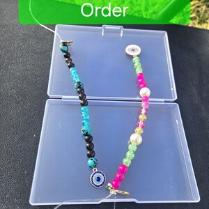 May include: Two beaded strands inside a clear plastic case. One strand features black and turquoise beads with a blue evil eye charm. The other strand has pink, green, and white beads with a circular charm. The text Custom Made To Order is visible.