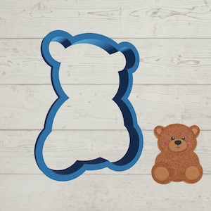 May include: Blue plastic cookie cutter shaped like a teddy bear. A brown teddy bear is sitting next to the cutter on a white wood background.