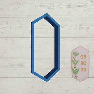 May include: A blue geometric cookie cutter in the shape of a hexagon with rounded corners. The cutter is on a white wood background. A pink and white cookie with green icing and the numbers 09 30 23 is in the background.