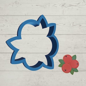 May include: Blue plastic cookie cutter shaped like a flower with three red and white holly berries with green leaves on a white wood background.