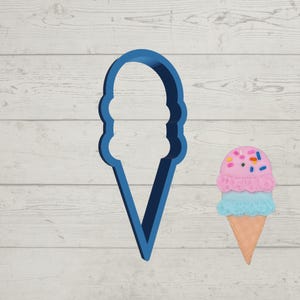 May include: A blue plastic ice cream cone cookie cutter next to a colorful ice cream cone. The ice cream cone has pink and blue scoops with sprinkles and a waffle cone base. The cookie cutter is a 3D outline of an ice cream cone.