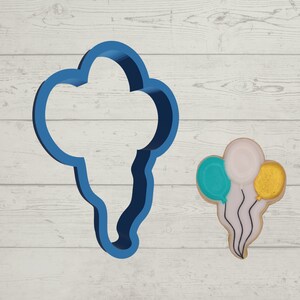 May include: A blue plastic cookie cutter shaped like a bunch of balloons. A decorated cookie with three balloons in the colors of teal, white, and gold is shown to the right.