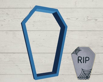 May include: A blue plastic cookie cutter shaped like a coffin. A gray cookie with a spiderweb design and the text "RIP" is next to the cutter.
