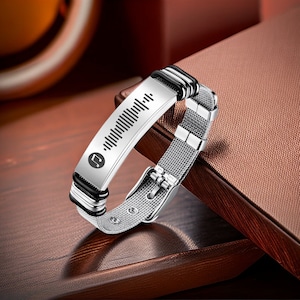 Può includere: Bracciale argentato con cinturino a rete e placca rettangolare con forma d'onda musicale e simbolo di nota musicale. Il bracciale ha dettagli neri e chiusura con fibbia. Presentato su una superficie marrone.