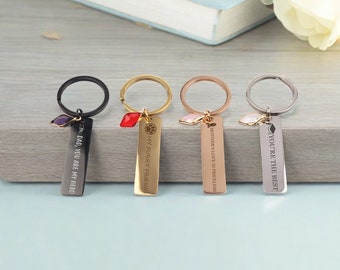 May include: Four metal keychains with engraved messages. The keychains have different colors: black, gold, rose gold, and silver. Each keychain has a rectangular tag with a message and a decorative charm. The messages include "DAD, YOU ARE MY HERO" and "YOU'RE THE BEST."