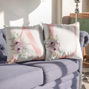 May include: Two white throw pillows with pink floral designs and large pink letters "A" and "U". The pillows have a watercolor style with pink flowers, green leaves, and yellow accents. They rest on a blue sofa.