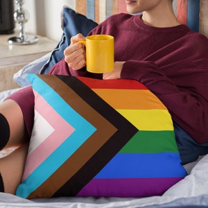 May include: A cushion displaying the Progress Pride flag design. The cushion has a white background with a pink, light blue, brown, black, and blue chevron, and a rainbow of colours. A yellow mug is held in the person's hand.