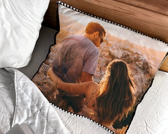 May include: Square cushion featuring a printed image of a couple embracing, set against a scenic backdrop. The cushion has a black border with white pom-pom trim. It rests on a white quilted bedspread, near a white pillow and wooden surface.