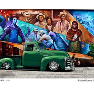 May include: A green vintage pickup truck parked in front of a colorful mural depicting people and a cityscape. The mural includes the words "I Pacific" and "Jordan Downs Copy Right 2025".