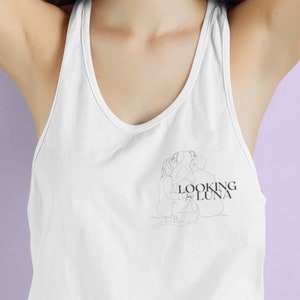 Puede incluir: Una mujer con el pelo largo y castaño lleva un top blanco de tirantes con un dibujo de línea negra de una mujer y el texto "LOOKING FOR LUNA" en la parte delantera.