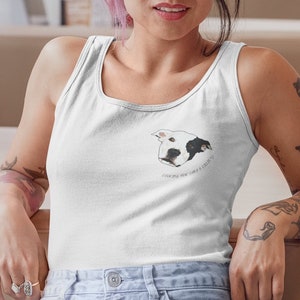 Puede incluir: Un top blanco de tirantes con una ilustración de un perro blanco y negro y el texto "Good things for a little boy". La modelo lleva el top y unos vaqueros azules.