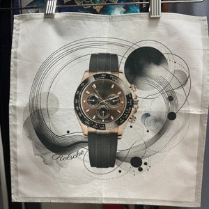 Puede incluir: Una toalla de té blanca con un reloj negro y dorado impreso en ella. El reloj tiene una correa negra y una esfera marrón con marcas blancas. El fondo es un diseño abstracto blanco y gris con círculos y líneas.