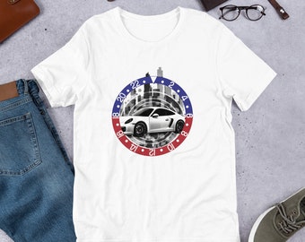 Puede incluir: Camiseta blanca con un gráfico de un coche deportivo blanco conduciendo por un horizonte urbano. El coche está rodeado por un círculo rojo, blanco y azul con los números del 18 al 22 y del 2 al 8.