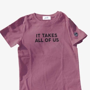 Peut inclure: T-shirt mauve avec l'inscription noire "IT TAKES ALL OF US". Un symbole de paix est sur la manche gauche. Le t-shirt a un col rond et une petite étiquette au niveau de l'encolure.