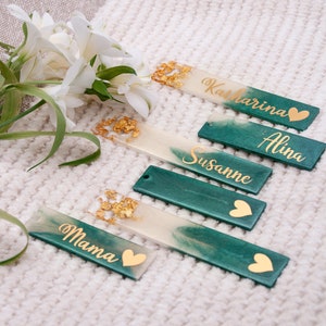 Könnte beinhalten: Fünf grüne und weiße Harz-Lesezeichen mit goldenen Blatt-Akzenten und goldenen Herzen. Die Lesezeichen sind mit den Namen "Mama", "Susanne", "Alina" und "Katharina" personalisiert.