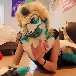 May include: A detailed close-up of a dinosaur-themed fursuit head with a tan, teal, and white color scheme. The fursuit features large, expressive eyes and a detailed snout. The image captures the intricate design and craftsmanship of the costume.
