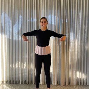 May include: A person wearing a black long-sleeved top and black leggings, with a light pink heating pad around the waist. The pad is segmented.