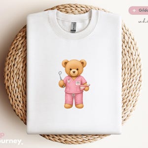 May include: White sweatshirt with a teddy bear dentist illustration. The bear, in pink scrubs, holds dental tools. The sweatshirt is a Gildan 18000, displayed on a woven, circular surface. The design is a watercolor style.