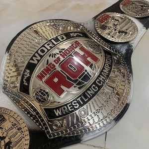 May include: A wrestling championship belt with a silver and black design. The belt features the text "WORLD RING OF HONOR WRESTLING CHAMPION" in red and black. Additional smaller medallions are also visible.