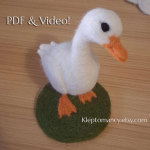 Puede incluir: Un ganso de fieltro blanco hecho a mano con pico y patas naranjas, posado sobre una base de fieltro verde. El texto "PDF & Video!" está encima del ganso, y el nombre de la tienda Etsy está debajo.