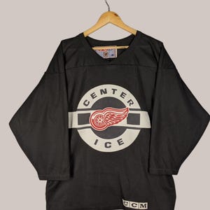 Puede incluir: Camiseta de hockey negra con un logotipo circular blanco que presenta una rueda alada roja y el texto "CENTER ICE". El logotipo está sobre un fondo rayado en blanco y negro. La camiseta tiene cuello en V y el texto "CCM" está impreso en la parte inferior.