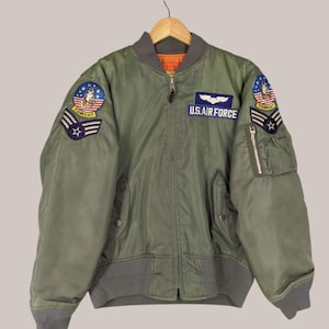 May include: A green bomber jacket with a zip fastening and two pockets. The jacket has three patches on the front: one with the words "U.S. AIR FORCE" and two with the word "TOMCAT".