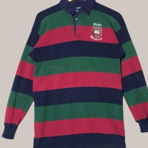 May include: A long-sleeved striped rugby shirt with a navy blue, green and red colour scheme. The shirt features an embroidered crest with the text "Oxford University".