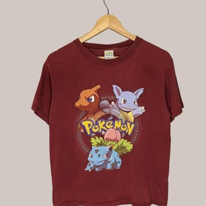 May include: A red t-shirt with a graphic of three Pokemon characters: Charmander, Bulbasaur, and Squirtle. The text "Gotta Catch 'Em All" is printed in a circle around the characters.