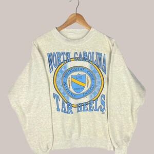 Op de afbeelding: Een lichtgrijs sweatshirt met een ronde hals en lange mouwen. De voorkant heeft een cirkelvormig embleem met de woorden "North Carolina" en "Tar Heels" in blauw, rond een schild met de Latijnse tekst "Universitas Carolina Septentrionalis".
