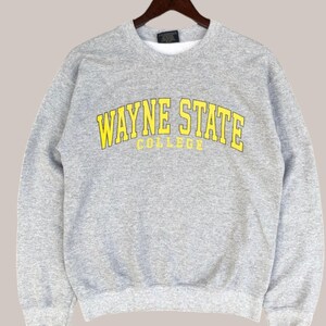 May include: A heather gray sweatshirt with the words "WAYNE STATE COLLEGE" in yellow block letters. The sweatshirt is hanging on a wooden hanger. The sweatshirt has a crew neck and long sleeves.