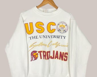 May include: White crewneck sweatshirt featuring the text "USC THE UNIVERSITY Southern California TROJANS" in yellow and maroon. The sweatshirt also has a university seal. The sweatshirt is hanging on a wooden hanger.