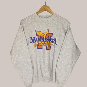 May include: A heather gray sweatshirt with the text "MINNESOTA STATE" in purple and orange, and a yellow eagle graphic. The sweatshirt is hanging on a wooden hanger.